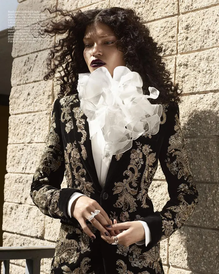 Femme en costume noir brodé, devant un mur de pierre, regardant l’appareil.
