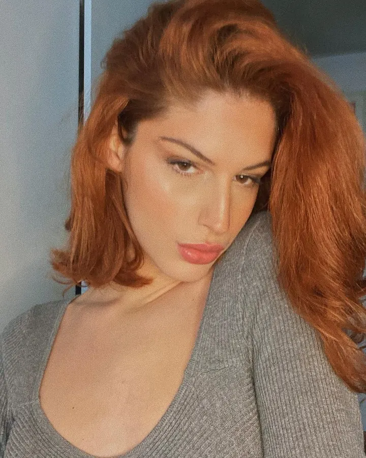 Femme aux cheveux roux, regard intense, vêtue d’un pull gris, légèrement penchée vers l’avant