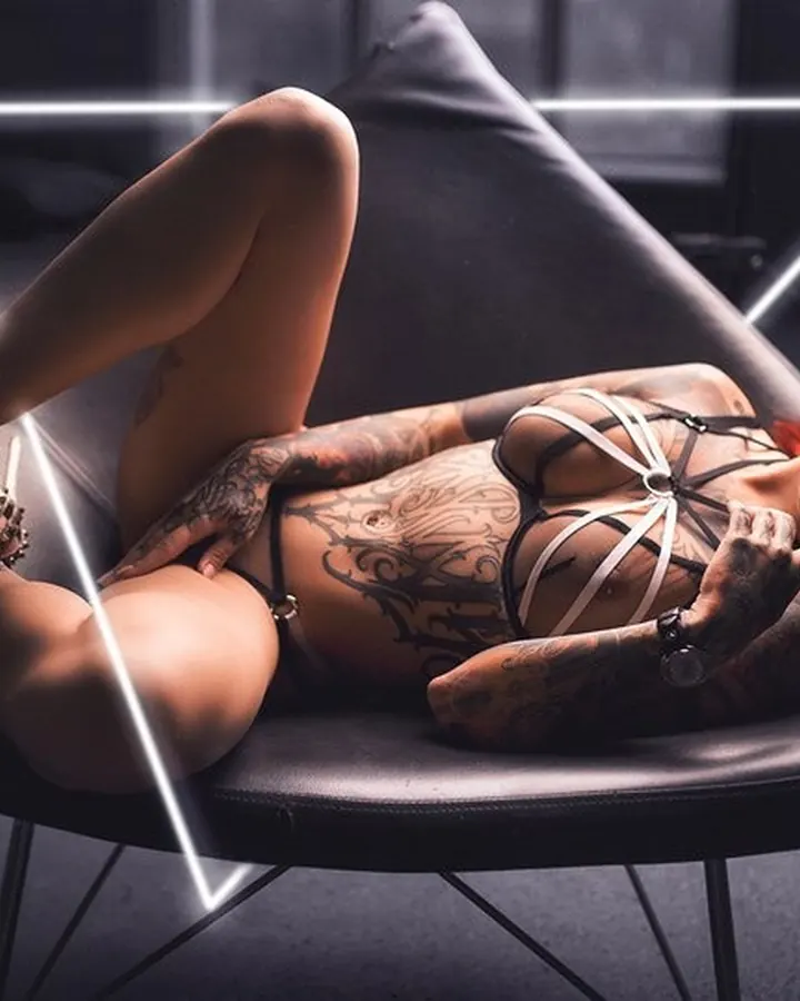Femme aux cheveux roux, en lingerie noire, allongée sur un fauteuil design, corps tatoué, regardant l'objectif.