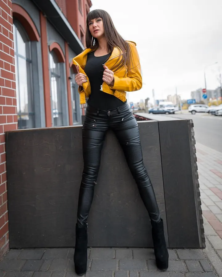 Femme aux cheveux bruns foncés, portant une veste jaune, un haut noir et un pantalon en cuir noir, posant en ville.