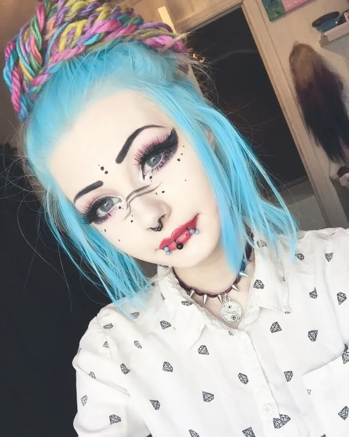 Femme aux cheveux bleus, maquillage exagéré, portant une chemise blanche avec des motifs de diamants, un collier avec un pendentif yin-yang.