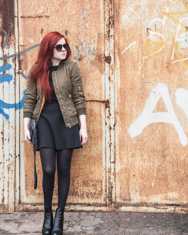 Femme aux cheveux rouges, vêtue d'une veste verte, jupe noire, collants noirs et bottes noires, debout devant un mur rouillé avec graffitis.