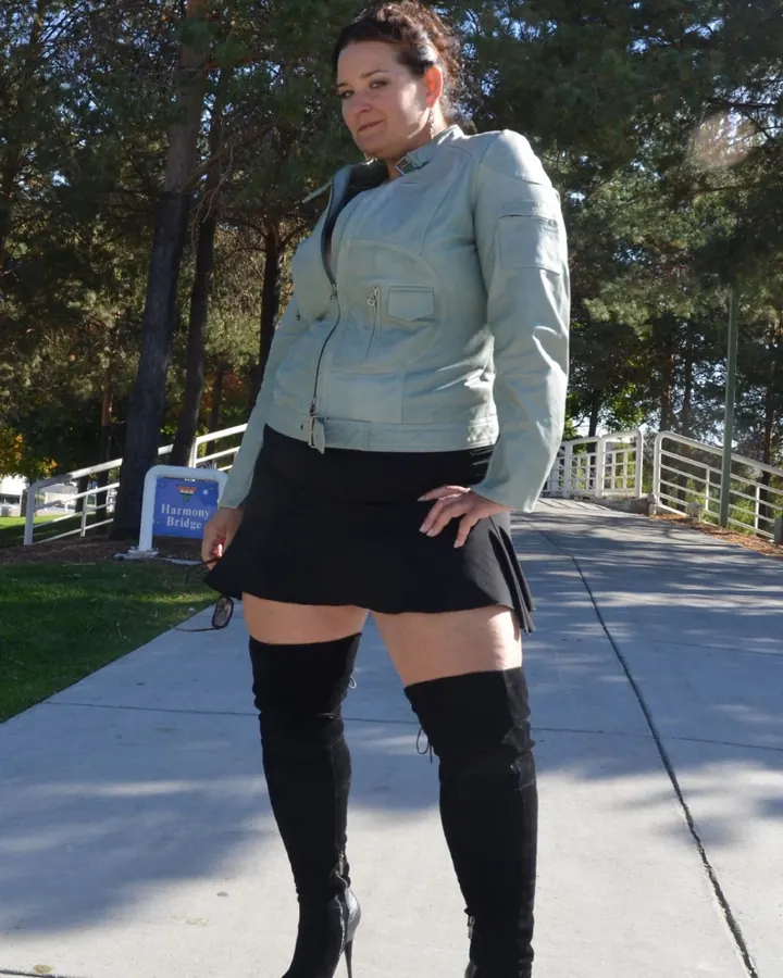 Femme en veste légère grise, jupe noire courte et bottes noires hautes, debout sur un trottoir ensoleillé près d'un pont.