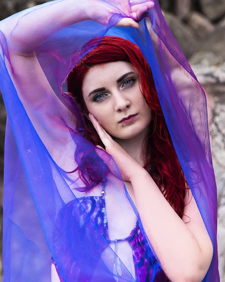 Femme aux cheveux roux et yeux bleus, portant un voile violet translucide, tenant son visage avec une main, regard intense.